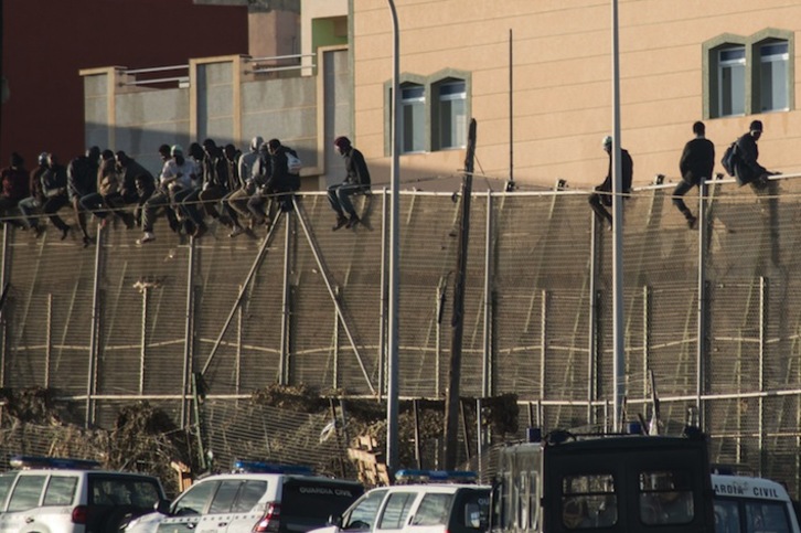 Un grupo de migrantes encaramados a la valla, con las furgonetas policiales abajo. (Blasco de AVELLANEDA/AFP) 
