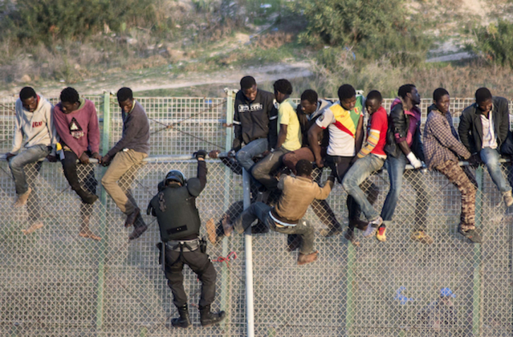 Varios migrantes encaramados a la valla de Melilla, mientras un agente policial intenta subir. (Blasco AVELLANEDA/AFP)