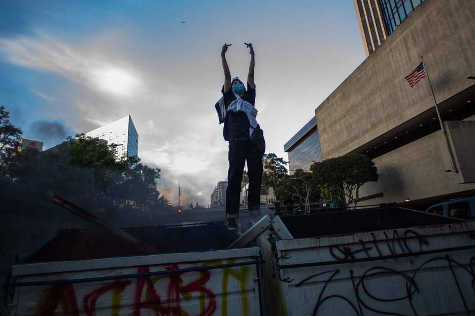 Un hombre encima de cubos de basura en llamas realiza gestos contra la policía en el centro de Los Ángeles. (Apu GOMES/AFP)