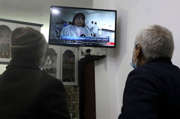 Dos palestinos ven en la televisión a la ministra de Salud palestina, Mai al-Kaila, anunciando el inicio de la vacunación contra el covid-19 en Cisjordania. (Hazem BADER/AFP)