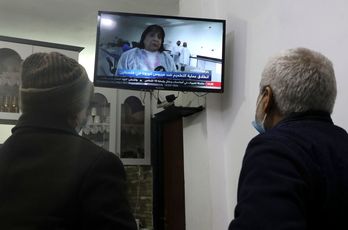 Dos palestinos ven en la televisión a la ministra de Salud palestina Mai al-Kaila anunciando el inicio de la vacunación contra Covid-19, en Cisjordania. (Hazem BADER/AFP)