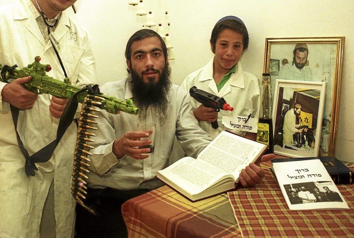 Gilad Sade (primero por la derecha) posa entre su padrastro y el retrato de Baruj Goldstein, un médico judío que mató a 29 palestinos. Fotografía: Ilan Mizrahi