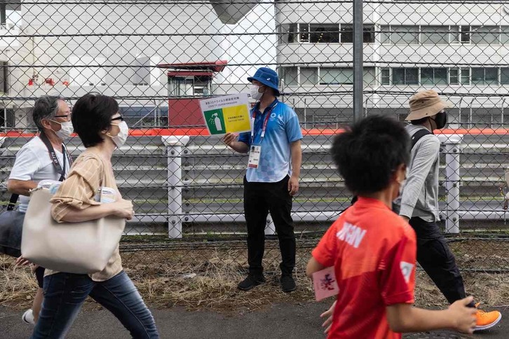 Olinpiar Jokoetako langile bat koronabirusaren aurkako neurriak hartzea gogorarazteko afixa batekin, zaleak txirrindularitzako lasterketa ikustera doazen bitartean. (Yuki IWAMURA/AFP)