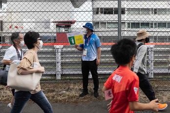 Olinpiar Jokoetako langile bat koronabirusaren aurkako neurriak hartzea gogorarazteko afixa batekin, zaleak txirrindularitzako lasterketa ikustera doazen bitartean. (Yuki IWAMURA/AFP)