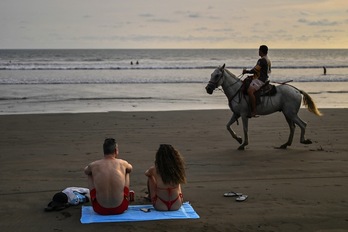 Turistas y un hombre que ofrece paseos a caballo por la playa de Jacó contemplan el mar.