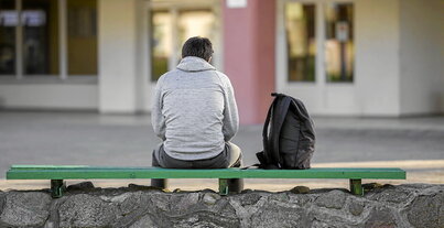 En la imagen de la izquierda, un joven sentado solo frente a un centro educativo. A la derecha, un grupo de alumnos en el aula, en una imagen de archivo.