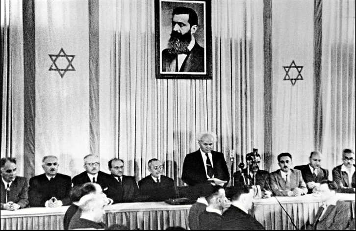 David Ben-Gurion lehen ministroa, Estatu israeldarraren sorreraren berri ematen, Juduen Nazio Kontseiluaren aurrean, eta Theodor Herzl sionismoaren sortzailearen irudia gainean duela.