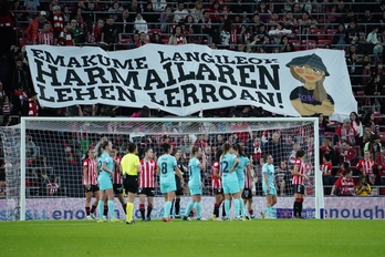 Jugadoras del Athletic y Barça se preparan para un saque de esquina, ante la pancarta conmemorativa del 8M.