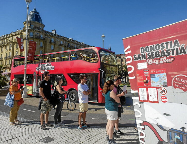 Euskal Herrira etorritako bisitari batzuk, eguzkipean, Donostiako autobus turistikoa hartzeko zain.