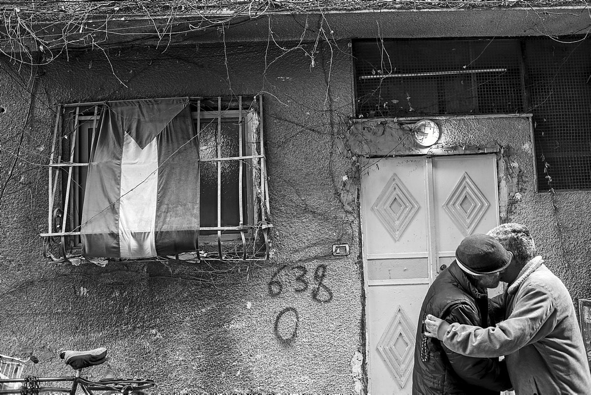 Dos hombres se abrazan en la calle frente a la bandera Palestina colgada en una ventana.