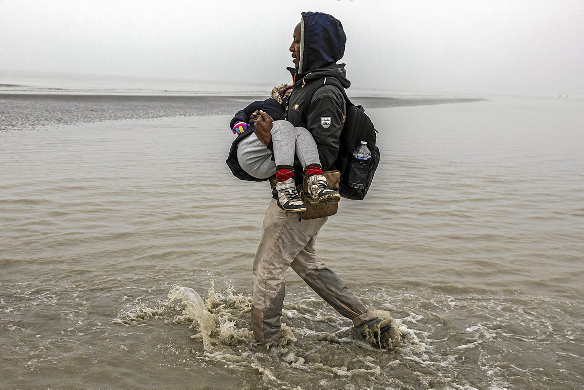 Un hombre procedente de Somalia lleva en brazos a su hijo hasta la embarcación que les llevará hacia Gran Bretaña. (Sergi Cámara)