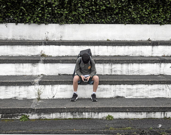 Un joven adolescente de Azkaine sentado en unos escalones él solo mientras pone toda su atención en el móvil que se encuentra entre sus manos.