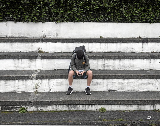 Un joven adolescente de Azkaine sentado en unos escalones él solo mientras pone toda su atención en el móvil que se encuentra entre sus manos.