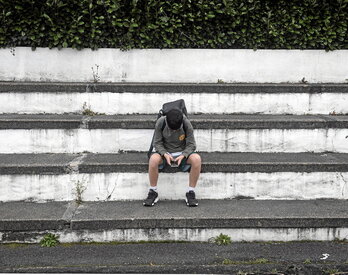 Un joven adolescente de Azkaine sentado en unos escalones él solo mientras pone toda su atención en el móvil que se encuentra entre sus manos.
