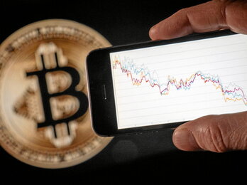 Una persona muestra en su smartphone la menguante cotización en bolsa del bitcoin, cuyo logotipo aparece al fondo de la imagen.