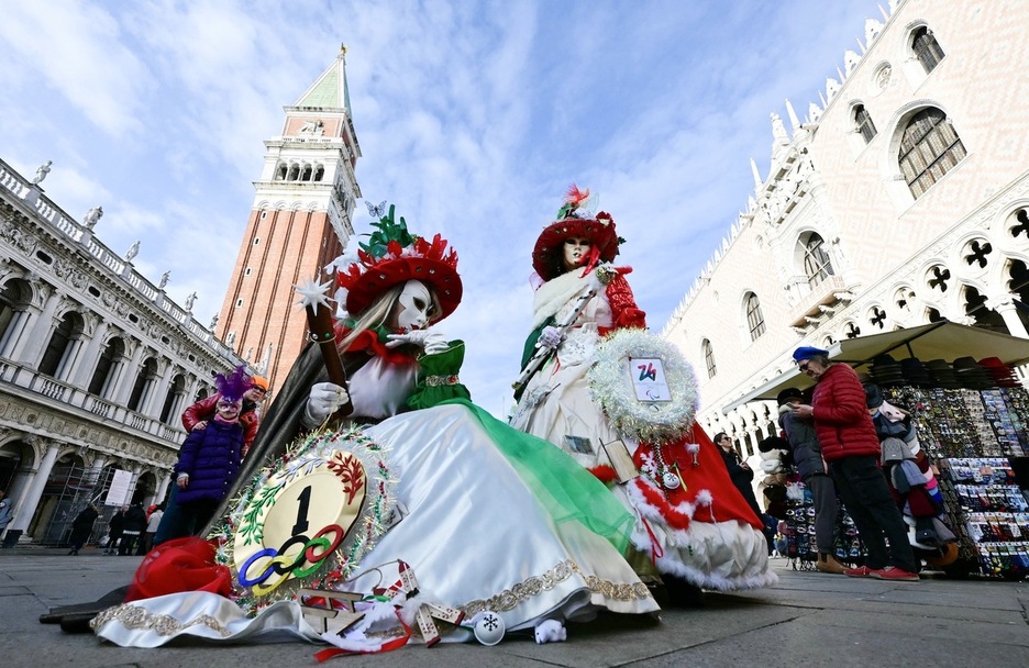 Dos personas vestidas con un disfraz con temática de los Juegos Olímpicos de Milán-Cortina 2026 y la bandera italiana posan en la plaza de San Marcos.