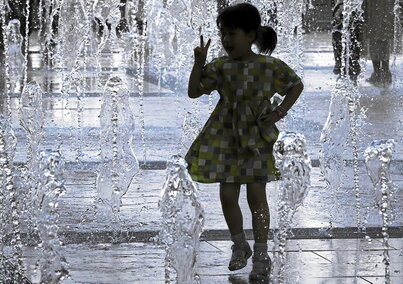 Behean, neskato bat iturri bateko urarekin jolasean, parke publiko batean. Eskuinean, eskolara bidean doan haur talde bat eta, azpian, ume bat patinete gainean errepide bat zeharkatzen.